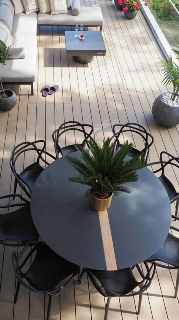 Pyöreä musta pöytä ja tuolit vaalean ruskealla terassilla. Terassin takaosassa näkyy terassikalusteita. Pöydän päällä on huonekasvi.